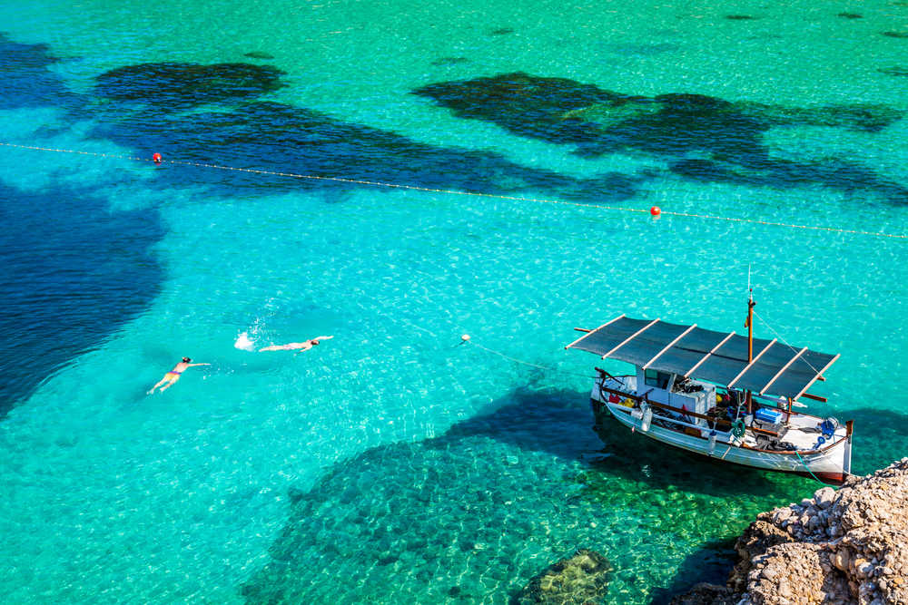 Vue en hauteur sur l'eau turquoise d'une plage d'Ibiza, on y voit un bateau et des personnes nager
