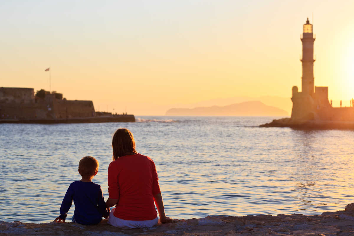 Une mère et son enfant regarde le soleil coucher sur les quais d'un port en Crète, un phare se tient au loin