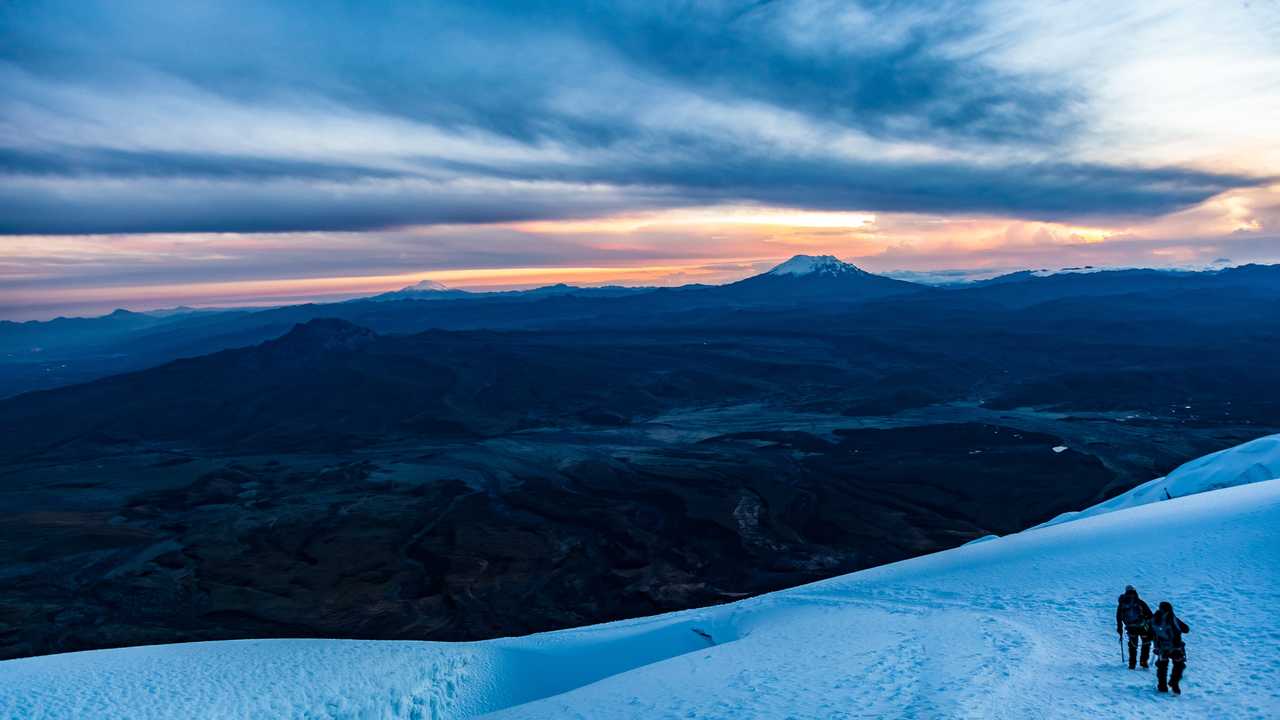 Deux alpinistes cordés marchent sur une crête enneigée à l'aube, avec une vue panoramique sur les montagnes et volcans d'Équateur sous un ciel de lever de soleil.