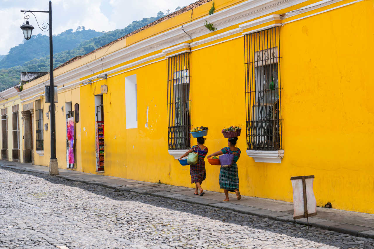 Deux femmes en tenues traditionnelles marchant avec des paniers de fruits sur la tête dans une rue pavée d'Antigua