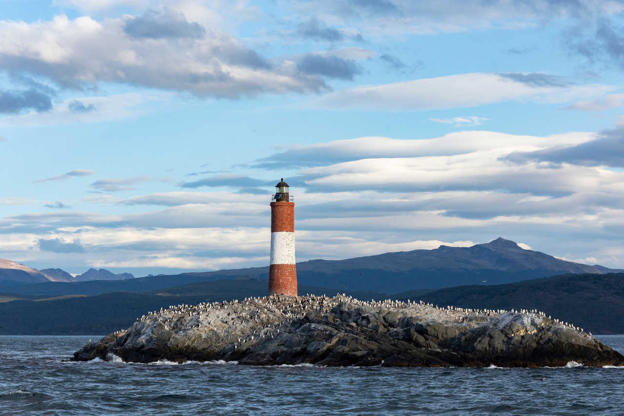 Le phare rouge et blanc Les Éclaireurs situé sur un îlot rocheux du canal de Beagle, dominé par les montagnes enneigées de la Terre de Feu au large d'Ushuaia, en Argentine.