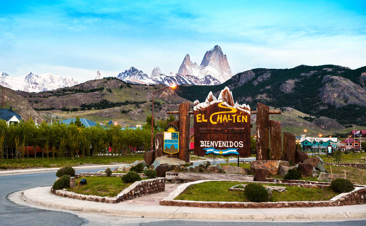 Panneau de bienvenue décoratif en bois à l'entrée du village d'El Chaltén, avec le village et les sommets enneigés et acérés du Fitz Roy et Cerro Torre en arrière-plan sous un ciel bleu en Patagonie argentine.