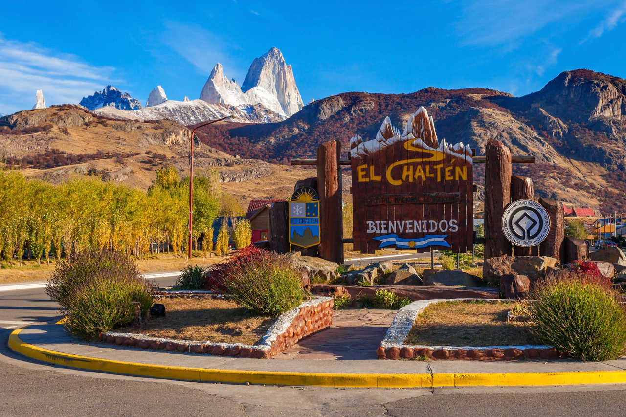 Panneau en bois de bienvenue à El Chaltén, capitale nationale du trekking, avec la route et le massif enneigé du Mont Fitz Roy en arrière-plan sous un ciel bleu en Patagonie argentine.
