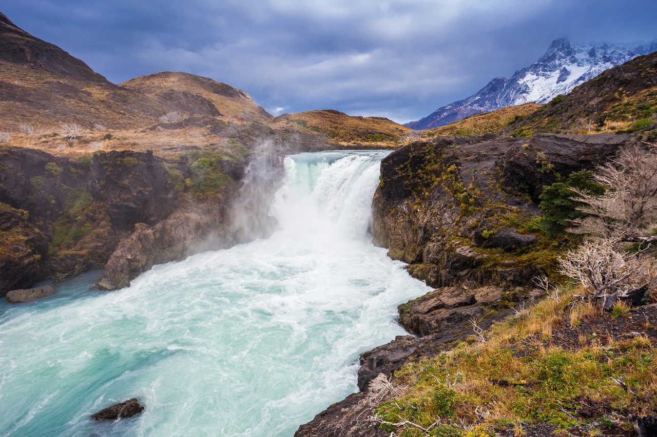 Puissante cascade aux eaux glaciaires turquoise de Salto Grande s'engouffrant dans une gorge rocheuse, avec les sommets enneigés du massif du Paine en arrière-plan, en Patagonie chilienne.