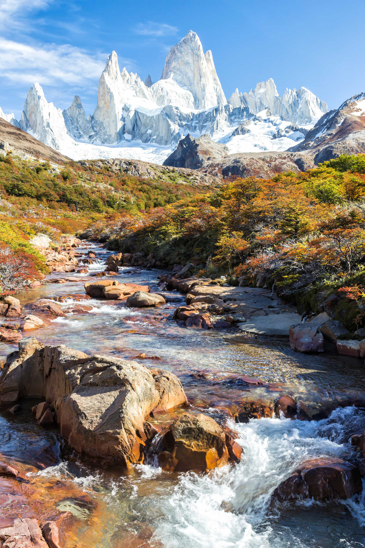 Rivière rocailleuse traversant une forêt aux couleurs d'automne rouges et orangées, dominée par les impressionnantes aiguilles granitiques et enneigées du Mont Fitz Roy en Patagonie argentine.