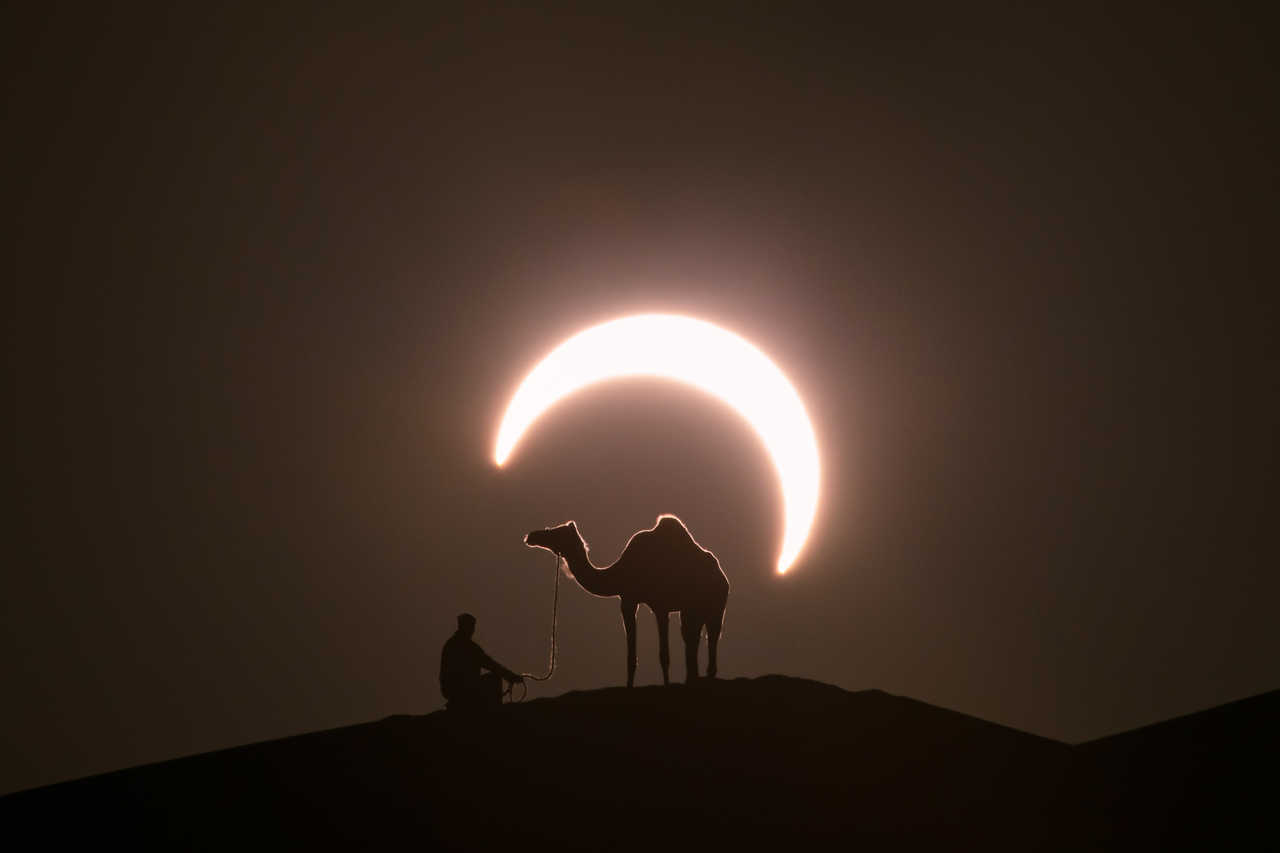 Silhouette d'un dromadaire et d'un homme sur une crête de dune de sable avec une éclipse solaire en arrière plan