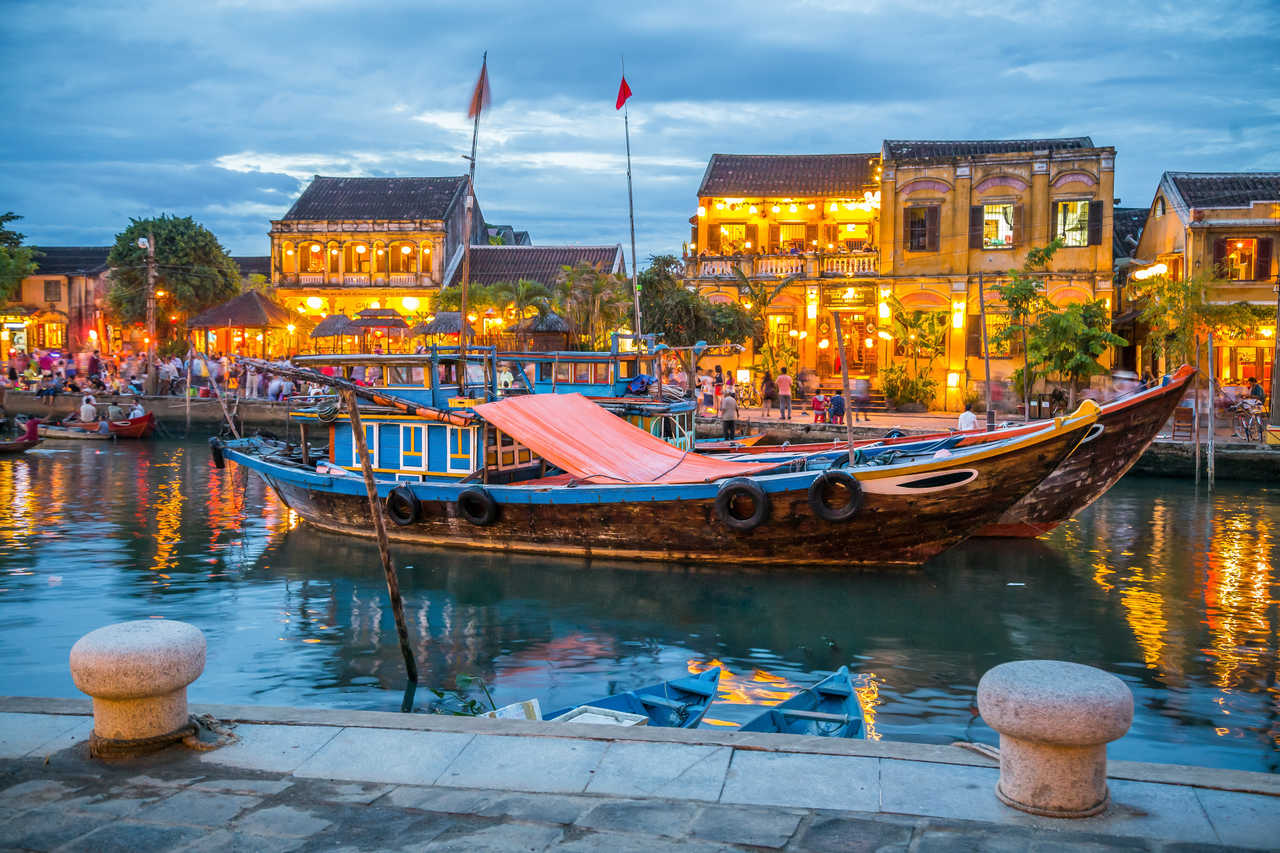 Un bateau fluvial en bois devant des bâtiments historiques illuminés au crépuscule à Hoi An, au Viêt Nam.