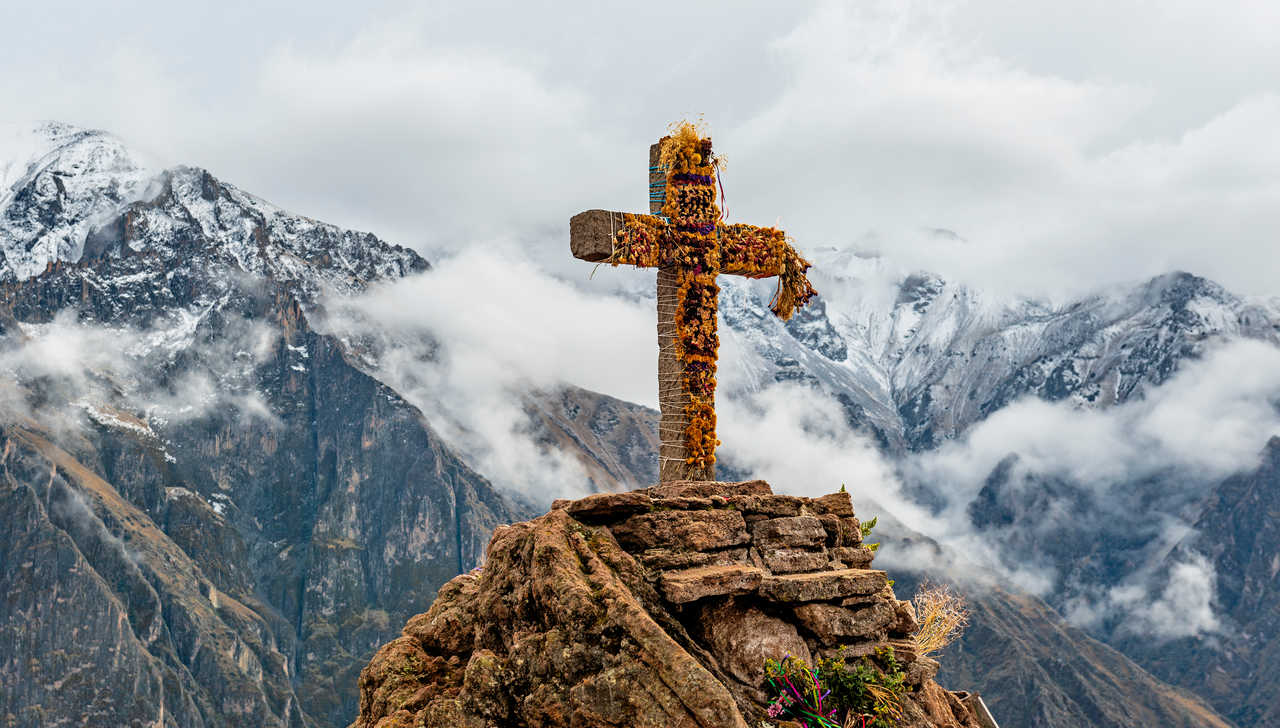 Une croix en pierre ornée de fleurs séchées se dresse au sommet d'un piton rocheux, avec en arrière-plan les sommets enneigés et les falaises abruptes du Canyon de Colca sous la brume.