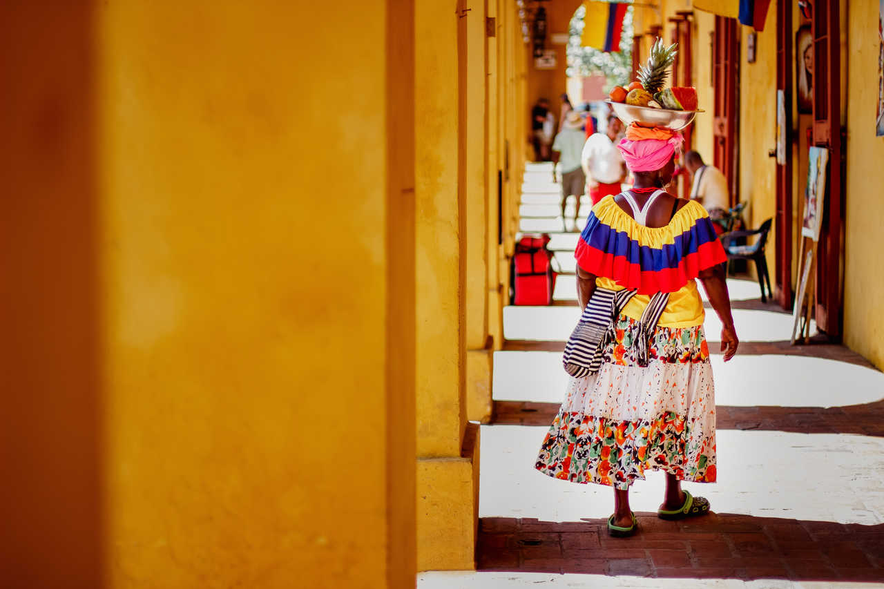 Une femme en tenue traditionnelle colorée portant un panier de fruits sur la tête, marchant sous les arches coloniales jaunes du centre historique de Carthagène des Indes.