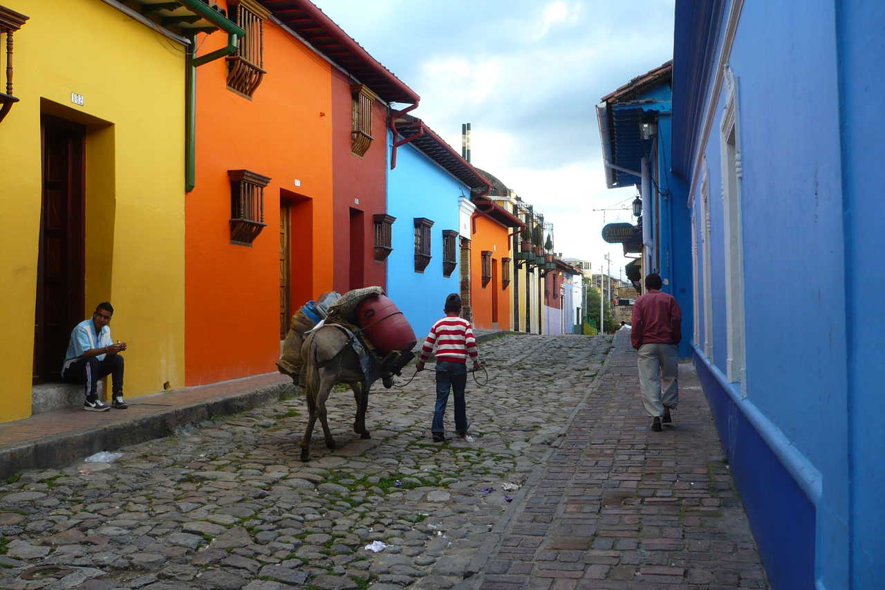 Une rue pavée pittoresque avec des maisons coloniales aux façades jaune, orange et bleu vif, où un enfant guide un âne chargé de marchandises à Bogota.