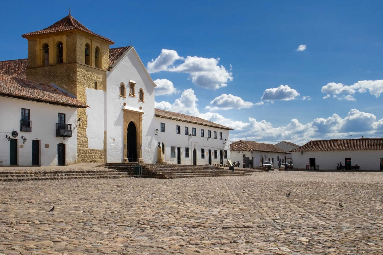 Vue panoramique de la grande place pavée de Villa de Leyva avec son église paroissiale blanche et ses bâtiments coloniaux sous un ciel bleu parsemé de nuages.
