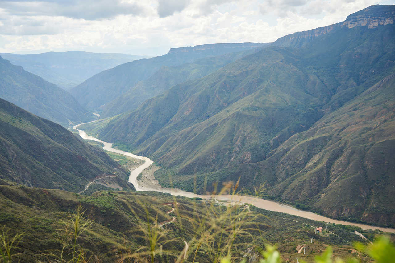 Vue panoramique spectaculaire sur les montagnes escarpées du Canyon de Chicamocha en Colombie, avec une rivière marron serpentant au fond de la vallée sous un ciel nuageux.