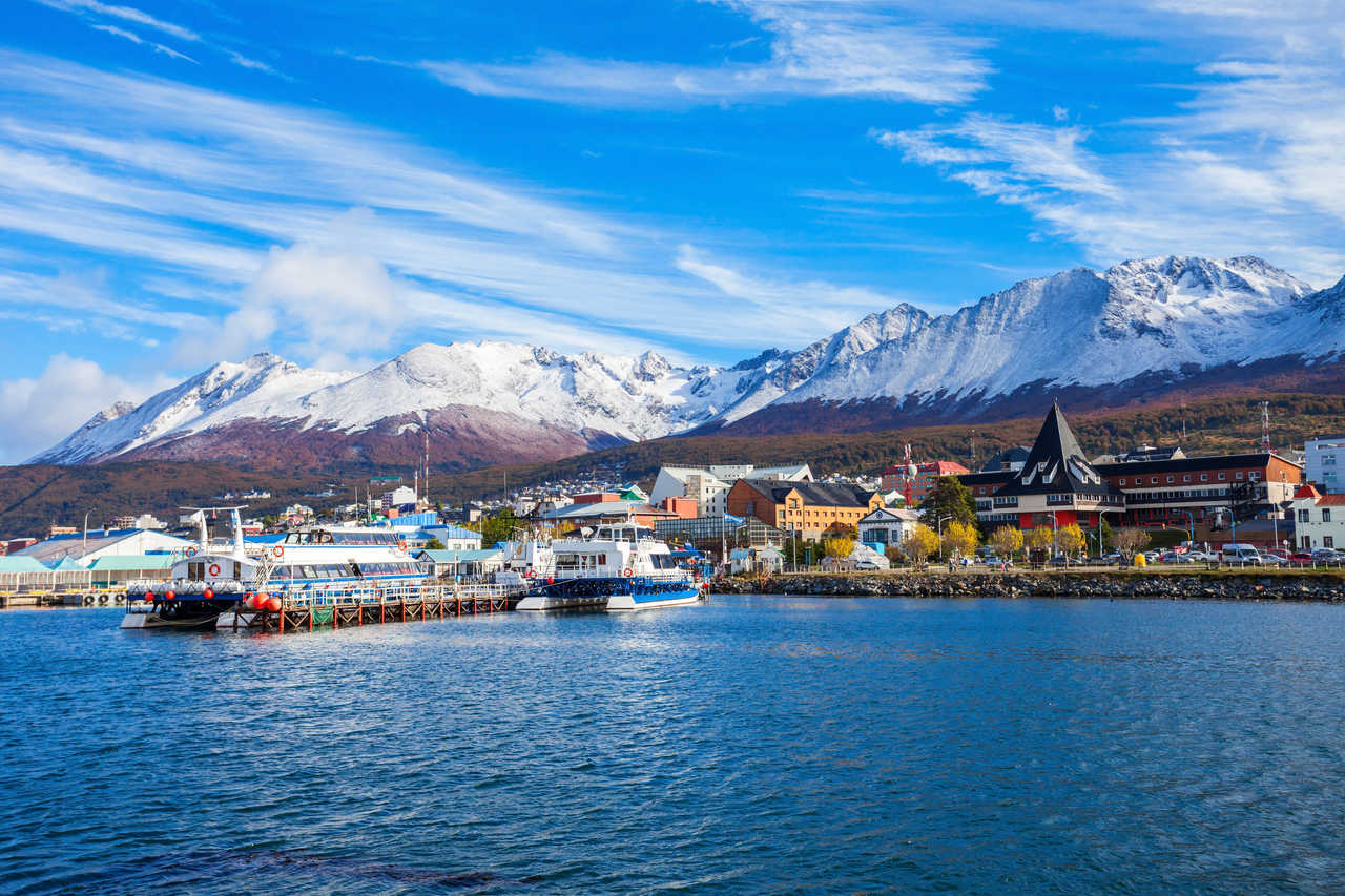 Vue panoramique sur le port d'Ushuaia avec bateaux à quai, la ville colorée et les sommets enneigés des Andes sous un ciel bleu en Patagonie argentine.