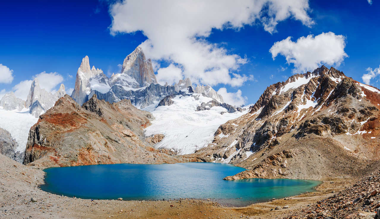 Vue panoramique sur les eaux turquoise du lac glaciaire Laguna de los Tres entouré de rochers, dominé par les glaciers et le sommet pointu du Mont Fitz Roy sous un ciel bleu, en Patagonie argentine.