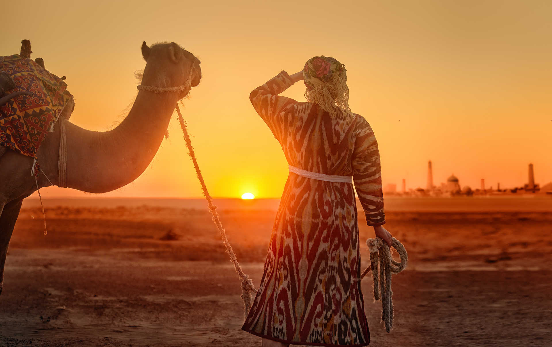 Une femme portant des vêtements nationaux traditionnels conduit un chameau dans le désert en direction de l'ancienne ville de Khiva au coucher du soleil.