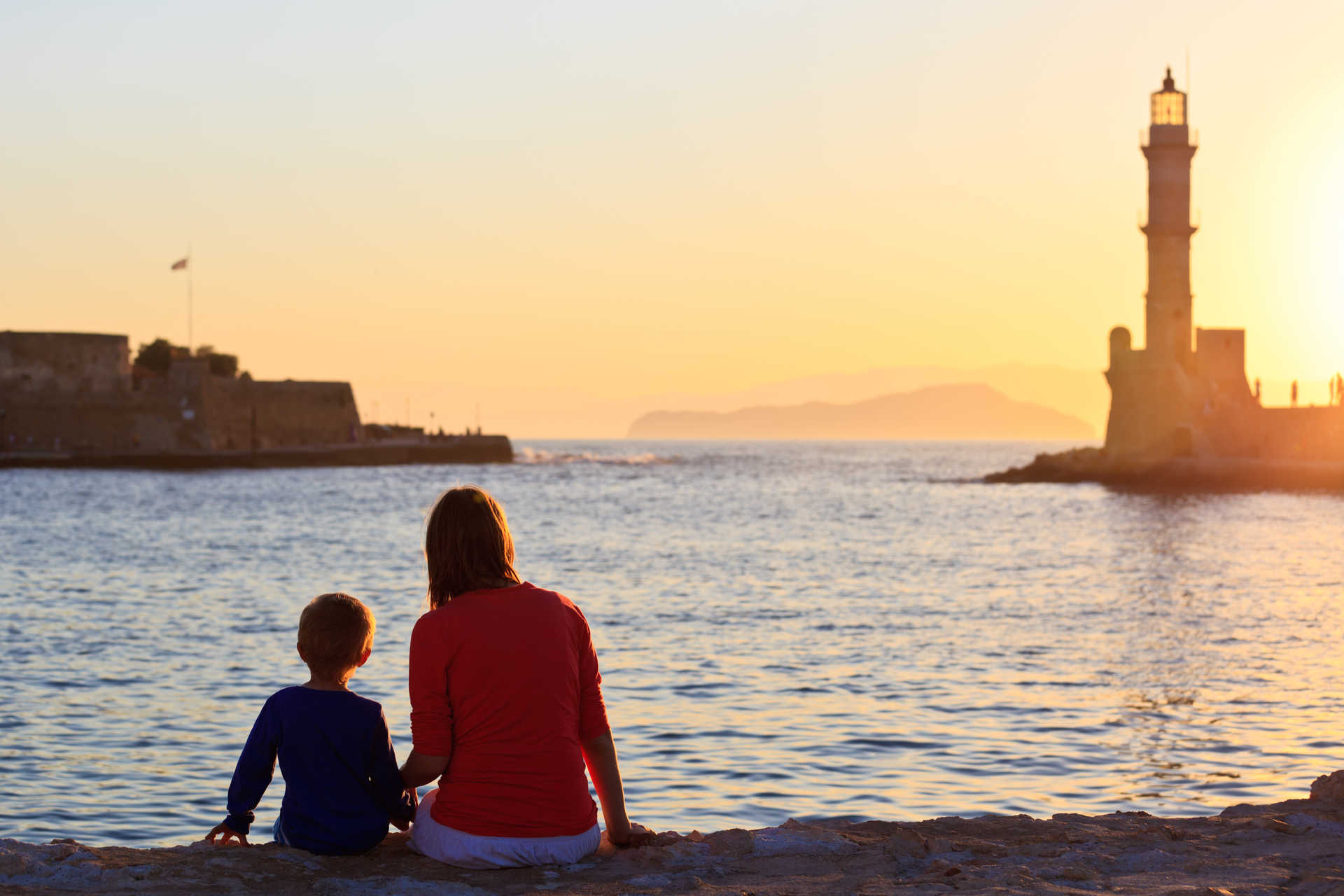 Une mère et son enfant regarde le soleil coucher sur les quais d'un port en Crète, un phare se tient au loin